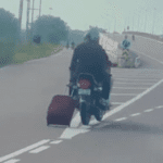 pillion-rider-pulls-trolley-bag-behind-transferring-bike-on-freeway,-web-surprised.-video